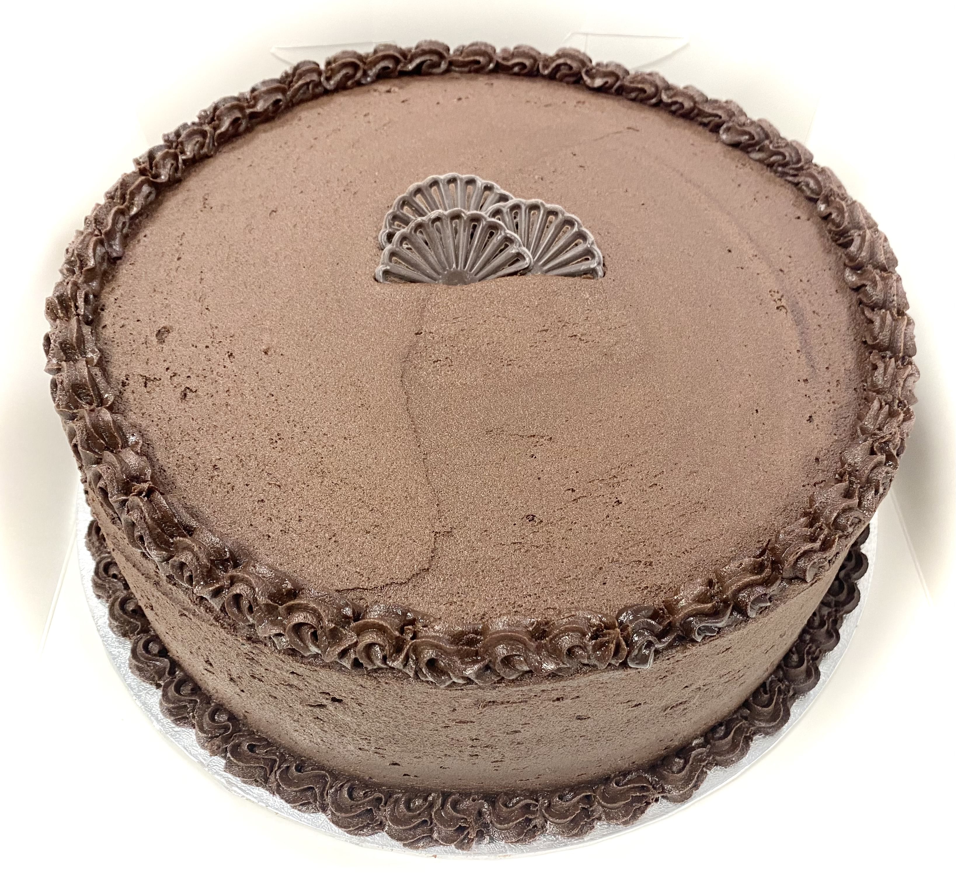 A round chocolate cake with smooth, decadent chocolate frosting, decorated with a border of swirled chocolate icing along the top edge. In the center, there is an embossed decorative fan-like design. This sumptuous dessert is placed on a white background.